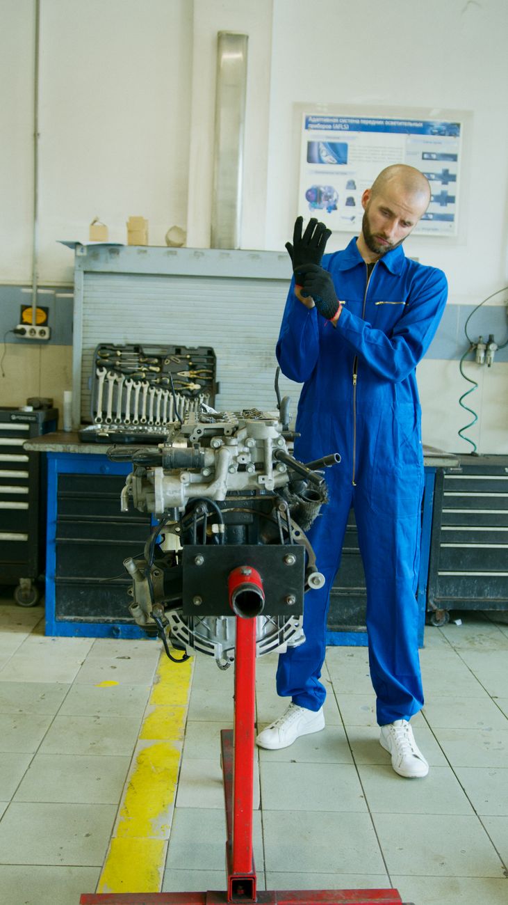 A mechanic in blue overalls adjusting gloves beside an engine in a garage